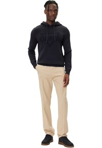 Felpa con cappuccio blu navy con un logo triangolare in rilievo, pantaloni beige e sneaker scure con suola marrone, in piedi davanti a uno sfondo bianco semplice.