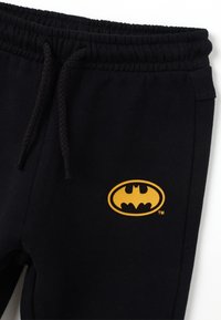 Zwarte katoenen sweatpants met een elastische tailleband, met een gele Batman-logo aan de zijkant. Zachte textuur en casual ontwerp.