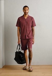 Bordo trumpai rankovėmis marškiniai ir jiems derančios šortai, pagaminti iš tekstūruotos medžiagos; modelis laiko juodą maišą ir dėvi kreminės spalvos šlepetes.