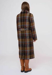Abrigo largo de cuadros con tonos marrones, dorados y azules, con un cuello estructurado y un detalle de correa en la parte posterior. Combinado con botas marrones hasta la rodilla.