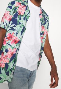 Chemise à manches courtes à boutons, fond bleu marine avec de grands motifs floraux roses et des feuilles vertes. Portée sur un T-shirt blanc.