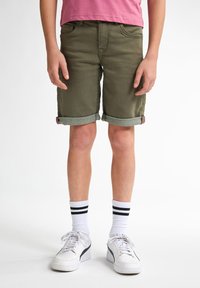 Pantalones cortos de mezclilla verde oliva con puños doblados, combinados con zapatillas blancas y calcetines a rayas negras. Modelo vistiendo una camiseta rosa.