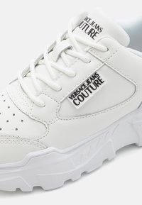 Baskets en cuir blanc avec des détails perforés, accents gris, semelle épaisse et étiquette logo noire affichant "VERSACE JEANS COUTURE."