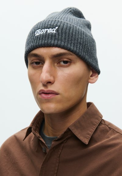 Giovane uomo che indossa un berretto grigio lavorato a maglia con la scritta "Bored" ricamata e una camicia marrone con bottoni, che guarda leggermente lontano dalla fotocamera.