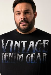 Camiseta de algodón negra con un gran texto desgastado en blanco y azul que dice "VINTAGE DENIM GEAR". Cuello redondo clásico.