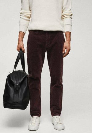 Mann trägt braune Hose und weiße Sneakers und hält eine schwarze Leder-Reisetasche an den Griffen vor einfarbigem Hintergrund.
