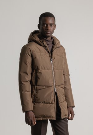 Chaqueta tipo puffer marrón con capucha, cierre de cremallera y bolsillos laterales. Tejido texturizado con diseño acolchado, llevada sobre un suéter de cuello alto marrón.