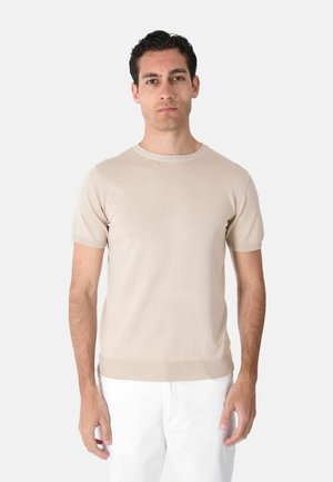 Uomo adulto con capelli scuri che indossa una maglietta beige a maniche corte in maglia e pantaloni bianchi, in piedi davanti a uno sfondo grigio chiaro uniforme.