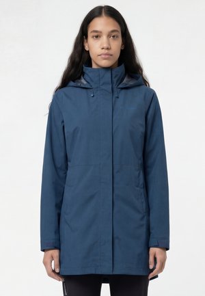 Jonge vrouw met lang donker haar, gekleed in een knielange marineblauwe waterdichte jas met een hoge kraag en kap, staand tegen een witte achtergrond.