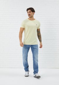 Light yellow cotton t-shirt with crew neck and small black logo, paired with blue jeans and black and white sneakers, against a white brick wall.
