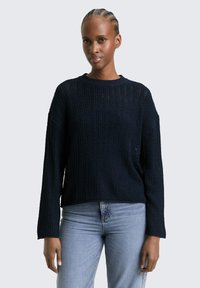 Vrouw met gevlochten haar, gekleed in een marineblauwe gebreide trui en lichtblauwe spijkerbroek, staand tegen een effen lichte achtergrond.