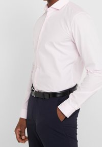 Camicia rosa chiaro a maniche lunghe con colletto a punta, apertura con bottoni e maniche con polsini. Abbinata a pantaloni scuri e una cintura nera.