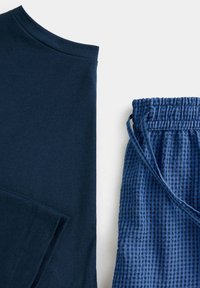 Chemise bleu foncé à manches longues à côté d'un pantalon à cordon bleu à carreaux sur fond blanc.