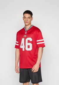 Piros NFL mez fehér 46-os számmal, hálós anyagból, fekete-fehér csíkos gallérral. Fekete rövidnadrággal viselve, sportos dizájnt és illeszkedést mutat.