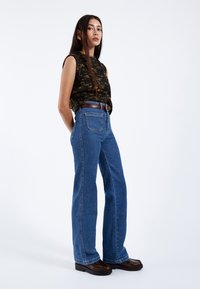 Jeans de tiro alto y pierna ancha en un denim azul medio con costuras en contraste, combinados con una blusa sin mangas de textura negra y un cinturón marrón.