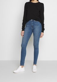 Blusa negra de manga larga con puños acanalados, combinada con jeans azul slim-fit y zapatillas blancas. Tela suave visible en la parte superior y en el denim.