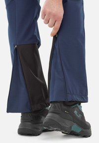 Pantalons d'extérieur bleus avec des aérations noires zippées sur les côtés ; tissu texturé, design fuselé. Des chaussures de randonnée noires avec des accents teal sont portées.