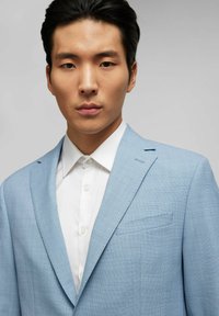 Lichtblauwe blazer van geweven stof, met een notch lapel en een enkele borstzak, gedragen over een witte overhemd met kraag.