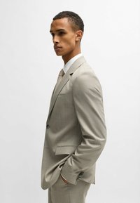Abito grigio chiaro con una sottile fantasia a scacchi; giacca monopetto, due pulsanti, rever stretti e vestibilità slim. Camicia bianca e cravatta strutturata.