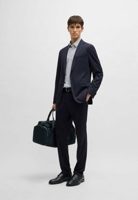 Abito blu navy con una vestibilità su misura, abbinato a una camicia leggera a fantasia. Gli accessori includono una borsa scura e scarpe in pelle nere.