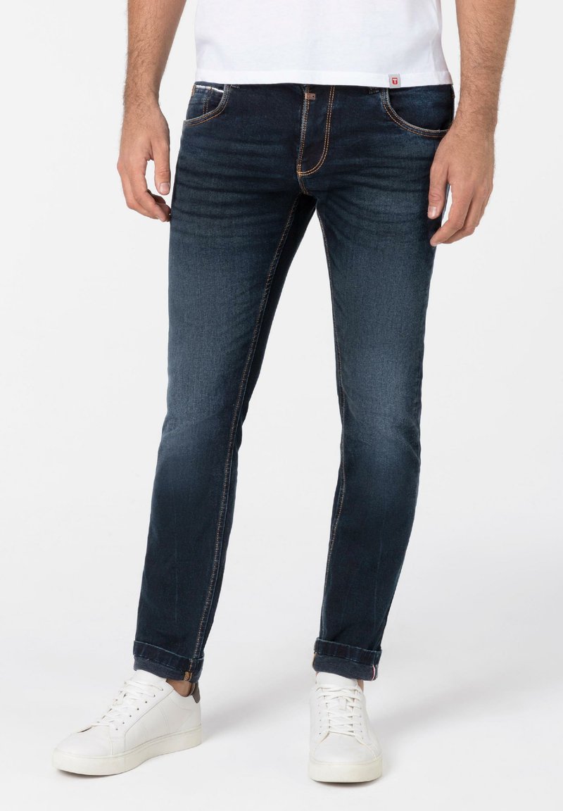 Dark blue jeans with a slim fit, featuring light blue stitching, five pockets, and a rolled hem, paired with white sneakers.