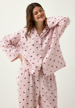 Kvinde med langt brunt hår, der smiler med lukkede øjne, iført lyserødt pyjamas med striber og små røde blomsterprint, hånd bag nakken.