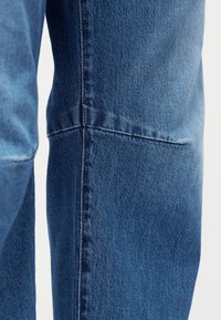 Κοντινή λήψη ποδιού μπλε τζιν, με έμφαση στη ενισχυμένη οριζόντια ραφή πάνω από την περιοχή του γονάτου.