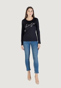 Donna con lunghi capelli castani che indossa una maglietta nera a maniche lunghe con "LuGo" in lettere lucide, abbinata a jeans blu e tacchi beige.