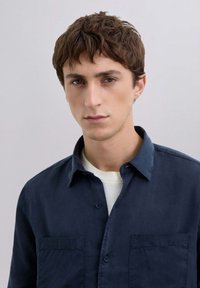 Camisa de botón azul marino con una textura suave y ligera, dos bolsillos en el pecho y un collar clásico, llevada sobre una camisa blanca.