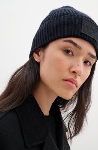 Black knitted beanie with a ribbed texture and a rectangular leather patch. The beanie snugly fits the head, featuring a dark color.
