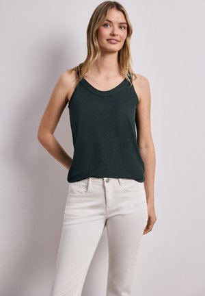 Młoda kobieta o blond włosach, ubrana w ciemnozielony bezrękawnik i białe spodnie, stoi na tle jednolitego jasnego tła.