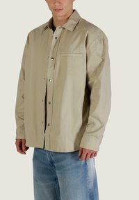 Camicia beige con bottoni, colletto appuntito, maniche lunghe e chiusure a scatto. Presenta una tasca sul petto e una trama sottile. Indossata sopra una maglia nera.