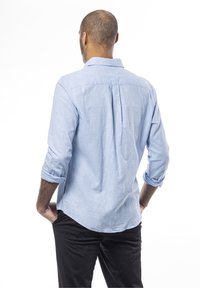 Hombre con las mangas remangadas, vestido con una camisa azul clara y pantalones negros, de pie de espaldas con las manos en los bolsillos frente a un fondo blanco.