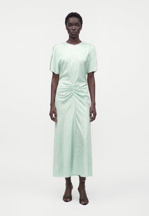 Femme grande aux cheveux courts portant une robe midi froncée vert menthe clair et des sandales à talons ouvertes noires, debout devant un fond uni.