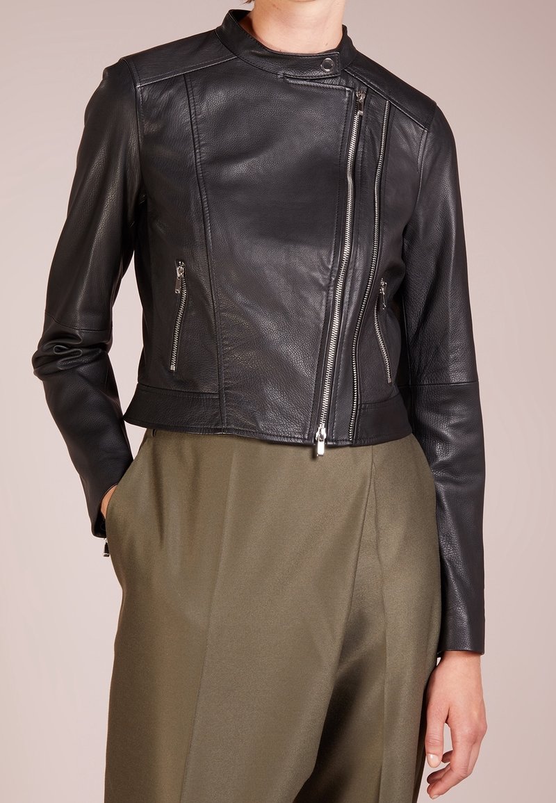 Femme portant une veste en cuir noire cintrée avec une fermeture éclair asymétrique à l'avant et des poches zippées, associée à un pantalon olive taille haute.