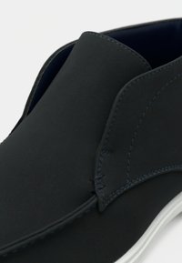 Chaussure noire avec une texture lisse et mate, présentant des coutures contrastantes bleu marine et un design arrondi. Semelle blanche, minimaliste et moderne.