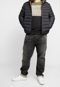 Giacca nera imbottita con cappuccio, maglione verde oliva e nero con motivo geometrico, jeans grigi e sneaker bianche.
