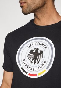Fekete pamut póló, amelyen kör alakú, nyomtatott mintázat látható, benne egy sas-emblémával, a "DEUTSCHER FUSSBALL-BUND" felirattal, valamint piros, sárga és fekete színű díszítésekkel.