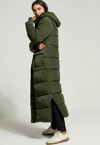 Lungo cappotto puffer verde con cappuccio, caratterizzato da un design trapuntato, apertura laterale con zip e tasche laterali. Indossato sopra pantaloni neri e sneakers.