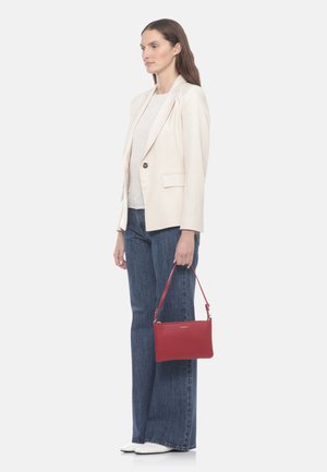 Donna in piedi di profilo che indossa un blazer color crema, una maglietta bianca, jeans a gamba larga, scarpe bianche, che tiene una borsa rossa per la cinghia con la mano destra.
