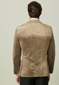 Brauner Wildleder-Blazer mit taillierter Passform, der eine glatte Textur und einen subtilen Glanz aufweist. Einzelner Schlitz hinten, kombiniert mit schwarzen Hosen.
