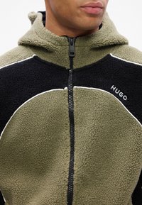Fleece bunda olivově zelené a černé barvy s předním zipem, bílými akcenty a vyšitým názvem značky "HUGO" na hrudi.
