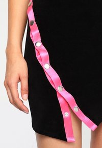 Svart klänning med sned nederkant, med en rosa satinkant och metallknappar längs ena sidan. Slät textur, figurnära design.