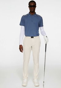 Golfasu, johon kuuluu vaaleansininen valkoisin raidoitus poolopaita, kermaisenväriset housut, valkoiset käsineet ja golfmaila. Sileä kangas ja räätälöity istuvuus.