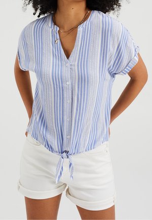 Femme portant une chemise à manches courtes à rayures verticales bleues et blanches nouée à la taille, avec un short blanc à ourlet roulé.