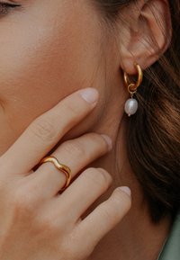 Prim-plan cu o mână care atinge fața, purtând un inel din aur ondulat și o cercea de aur în formă de cerc, cu o perlă albă atârnândă.