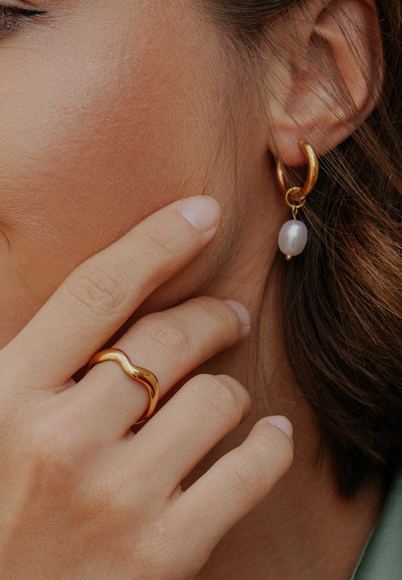 Prim-plan cu o mână care atinge fața, purtând un inel din aur ondulat și o cercea de aur în formă de cerc, cu o perlă albă atârnândă.