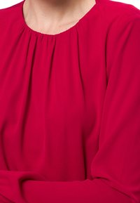 Blouse rouge en tissu doux et fluide, dotée d'un col froncé et de manches longues. La texture semble lisse et légère.