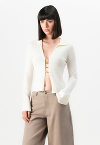 Langermet, hvit strikket topp med V-hals, med tre gullfargede klips foran, og en lett cropped hem. Kombinert med beige bukser.