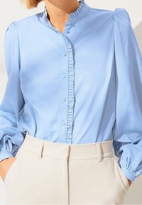 Blusa azzurra chiara con apertura a bottoni e colletto arricciato. Presenta maniche a sbuffo e tessuto morbido. Abbinata a pantaloni beige su misura.